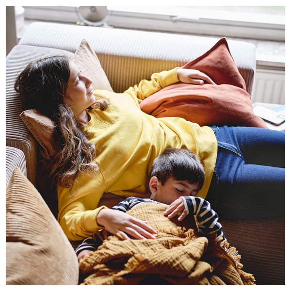 Una donna e un bambino in pigiama a righe giacciono su un divano sotto una coperta ruvida, creando una scena accogliente.