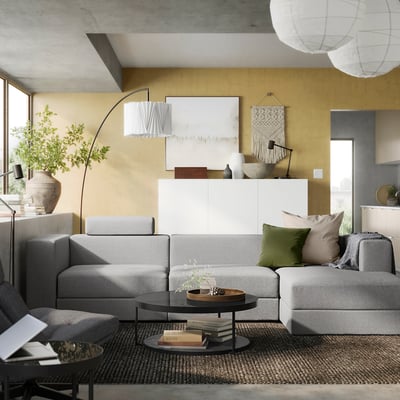 Soggiorno moderno con divano modulare JÄTTEBO grigio, cuscini verdi, tavolino in legno e pareti giallo senape, caratterizzato da un design contemporaneo.