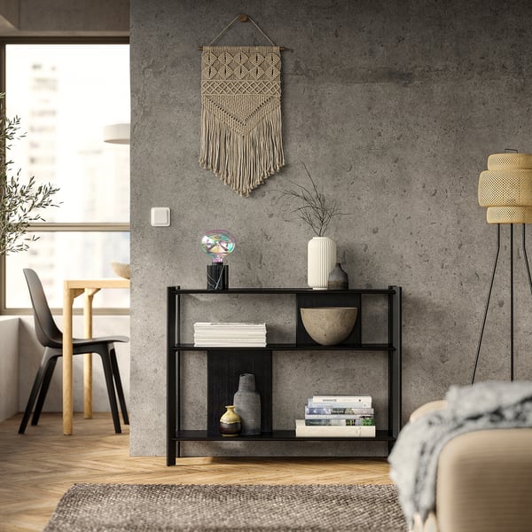 Soggiorno moderno con tavolino JÄTTESTA in bambù nero, libri decorativi, vasi e opere d'arte da appendere alle pareti. Divano beige chiaro e sedie in legno nelle vicinanze.