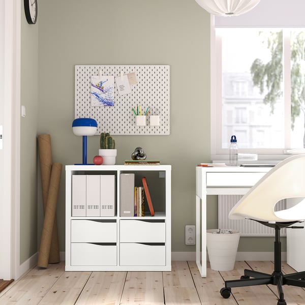 Scaffalature KALLAX con libri, piante, lampade e appunti. Scrivania bianca con computer e sedia vicino alla finestra.
