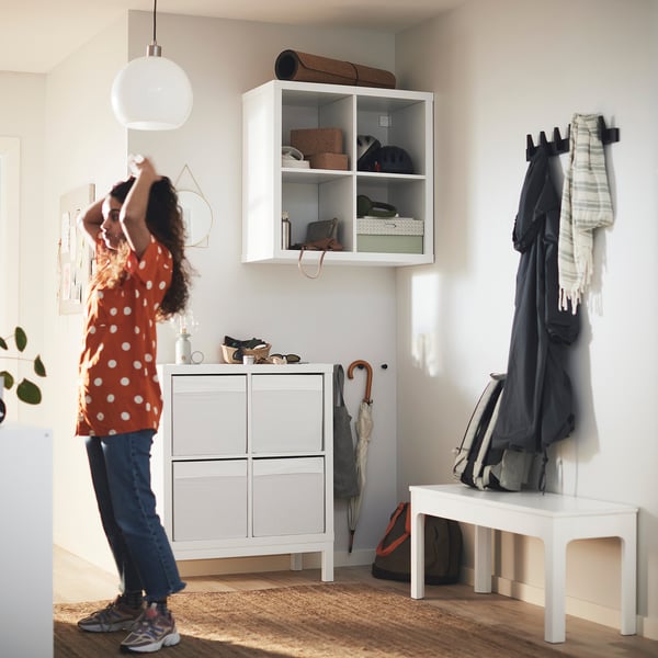 Una donna vicino agli scaffali KALLAX con gli oggetti. Appendiabiti e panca nelle vicinanze. Indossa una camicia rossa a pois.