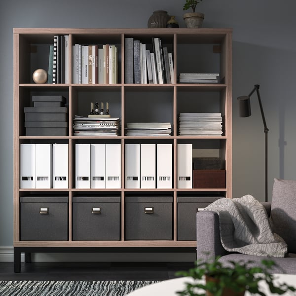 Libreria KALLAX in marrone chiaro, che ospita libri e raccoglitori. Adiacente a un'accogliente poltrona a motivi geometrici e pianta in vaso.