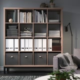 Libreria KALLAX in marrone chiaro, che ospita libri e raccoglitori. Adiacente a un'accogliente poltrona a motivi geometrici e pianta in vaso.