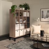 Mobile contenitore KALLAX in finitura legno, circondato da piante, libri e decorazioni per la casa, accanto a una sedia beige e un tavolo rotondo in vetro con set da caffè.