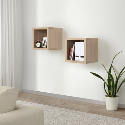 Mensole a muro KALLAX, legno chiaro. Piccoli cubi quadrati contengono libri e orologio. Accanto a una sedia beige e una pianta.