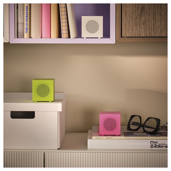 Tre colorati cubi di altoparlanti in verde, rosa e bianco su scaffali con libri e un paio di occhiali.