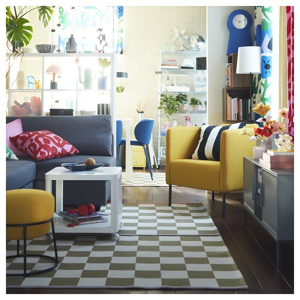 Luminoso soggiorno con divano grigio, poltrone gialle, tappeto a scacchi, tavolino bianco, decorazioni colorate e ampia finestra.