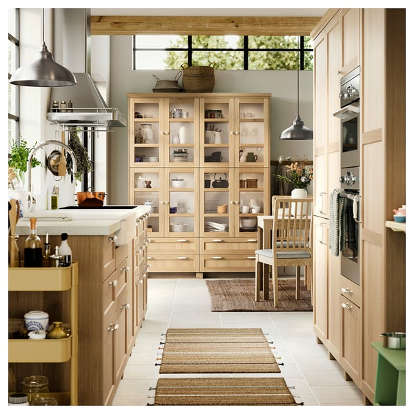Cucina moderna con armadi in legno chiaro, isola centrale, vetrina in vetro, lampade a sospensione e tappeti.
