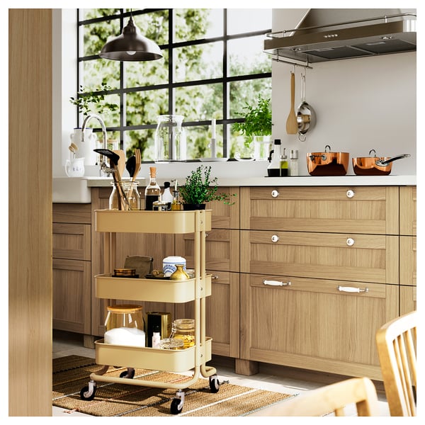 Cucina moderna con armadi in legno chiaro, un carrello giallo che contiene le forniture per cucinare e una grande finestra con verde all'esterno.