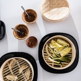 Vista dall'alto di un cestello per la cottura a vapore in bambù, piatti neri con verdure e gnocchi, ciotole di legno con salse e cucchiaio. Configurazione luminosa e pulita.