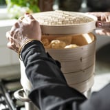 Persona che tiene il cesto a vapore in bambù con gnocchi, indossando un guanto nero.