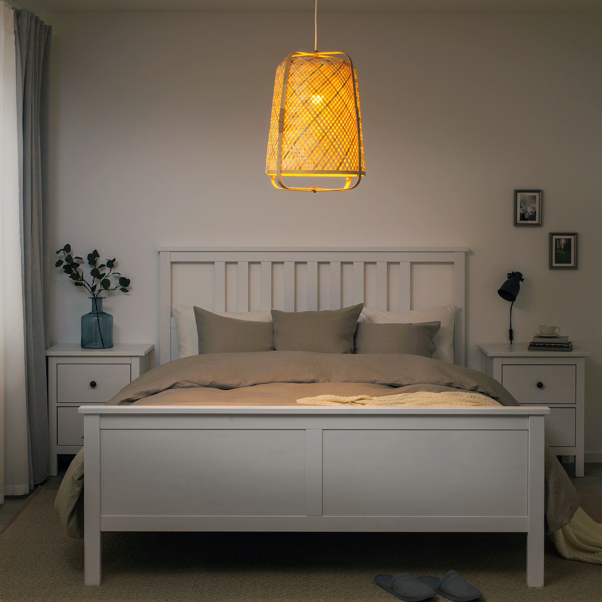 Camera da letto con lampada a sospensione KNIXHULT sopra il grande letto, la lampada è in bambù intrecciato, luce soffusa, il letto ha testiera in legno, due comodini con oggetti decorativi.