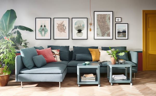 Soggiorno moderno con divano a forma di L verde acqua, cuscini colorati, due tavolini, piante e galleria d'arte a parete.