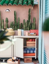 Piccolo mobile contenitore bianco con oggetti colorati e organizzati. Cactus in vaso in cima, barattoli blu, sacchetto rosso, scatole di legno all'interno.