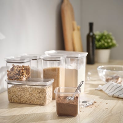 Cinque barattoli KORALLVIPPA conservano noci, cereali e polvere; trasparente, impilabile, con coperchi ermetici; Siediti su un tavolo di legno vicino al pane e alla pianta.