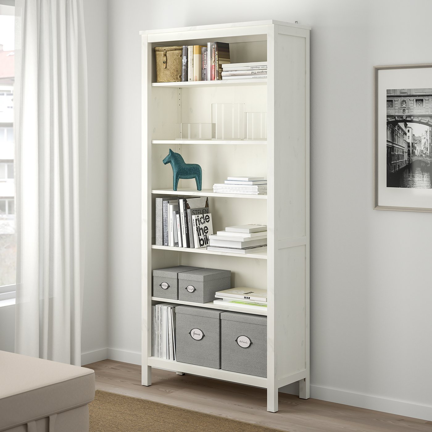 Scaffale KVARNVIK bianco sotto la finestra, che contiene libri, scatole e decorazioni.