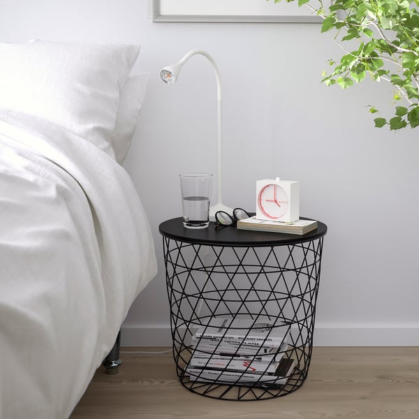 Tavolino in filo nero con motivo a rombi, orologio che regge, bicchiere d'acqua e lampada da lettura accanto al letto.