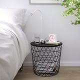 Tavolino in filo nero con motivo a rombi, orologio che regge, bicchiere d'acqua e lampada da lettura accanto al letto.