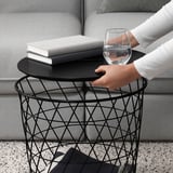Una persona che tiene in mano un moderno tavolino in metallo con un bicchiere d'acqua e libri, mostrando la sua portabilità e funzionalità di archiviazione.