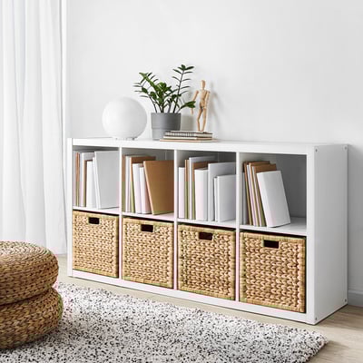 Scaffale kallax bianco pieno di libri e cesti intrecciati. La parte superiore contiene lampada, piante e decorazioni. I cestini conservano gli oggetti in modo ordinato.