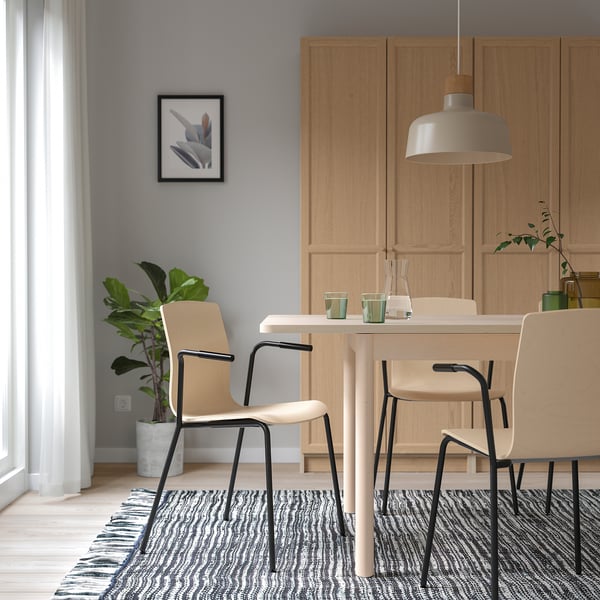 Moderna zona pranzo con tavolo in legno chiaro, sedie LÄKTARE in betulla, gambe in metallo nero, tappeto e lampada a sospensione. Pareti grigie, piante e arte.