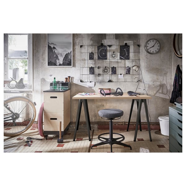 Postazione di lavoro in stile industriale con piano in legno e gambe in metallo nero, con vari oggetti decorativi e strumenti su un pavimento piastrellato.
