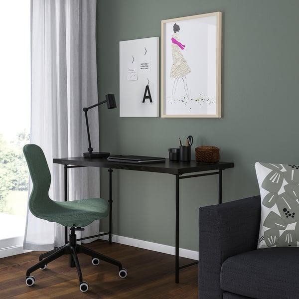 Home office con scrivania nera LAGKAPTEN. Sedia e lampada accanto. Il muro ha due cornici artistiche. Divano con un cuscino a destra.