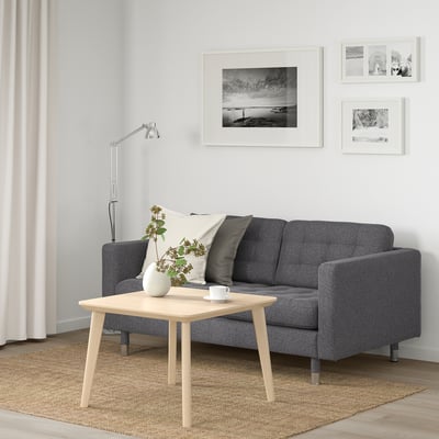 Accogliente soggiorno con divano grigio, tavolo in legno, lampada e foto. Cuscini bianchi e neri, fiori, tende leggere e tappeto beige.