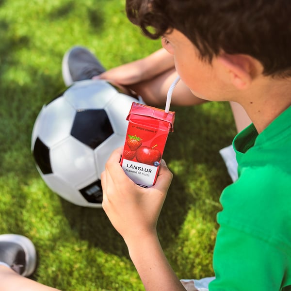Un bambino beve il frullato di frutta LÅNGLUR sull'erba vicino a un pallone da calcio, maglietta verde.