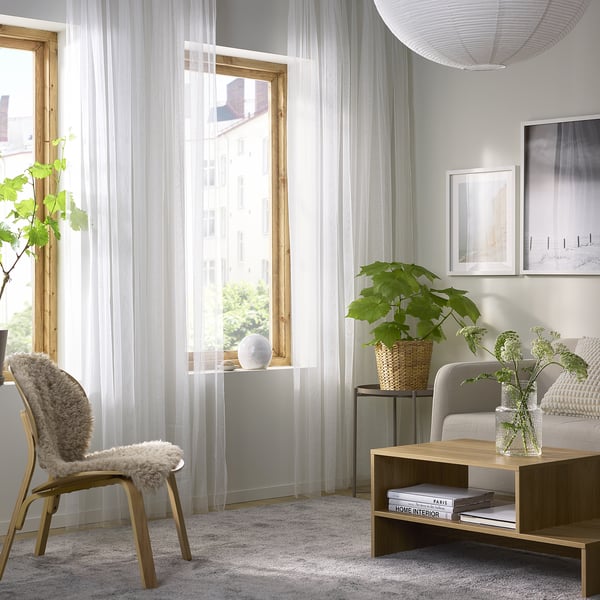 Soggiorno minimalista con grandi tende LILL trasparenti. Sedia in legno, tavolino e un divano con piante verdi e un tappeto bianco.