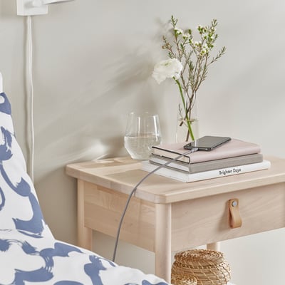 Un comodino con un cavo usb, fiori, un bicchiere e libri.