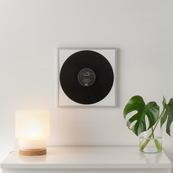 Un disco in vinile nero incorniciato era appeso a una parete grigio chiaro sopra uno scaffale bianco. La mensola contiene una piccola lampada e un vaso di vetro con foglie verdi.