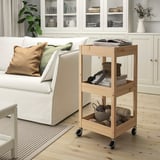 Carrello portaoggetti in legno accanto al divano, che contiene articoli per maglieria, libri e cestino. Il pino chiaro si abbina al divano bianco e al pavimento in legno.