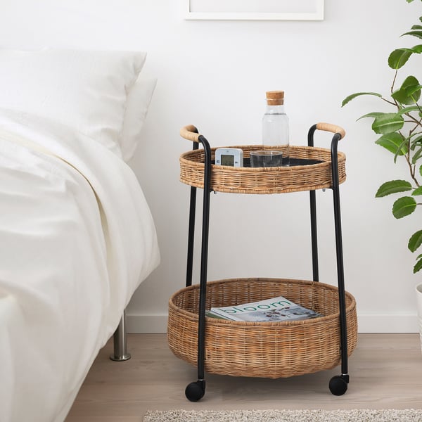 Carrello da comodino in rattan con due ripiani, che contiene orologio, bottiglia e rivista.