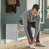 Uomo che lega scarpe da ginnastica bianche sulla panca MACKAPÄR, chinandosi con maglione grigio e pantaloni neri.