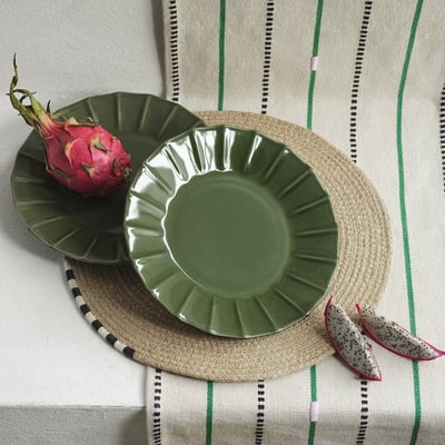 Il piatto in gres verde MÄVINN poggia su una tovaglietta rotonda intrecciata con frutto del drago. Il bordo smerlato dei piatti aggiunge consistenza, leggermente più grande della frutta.