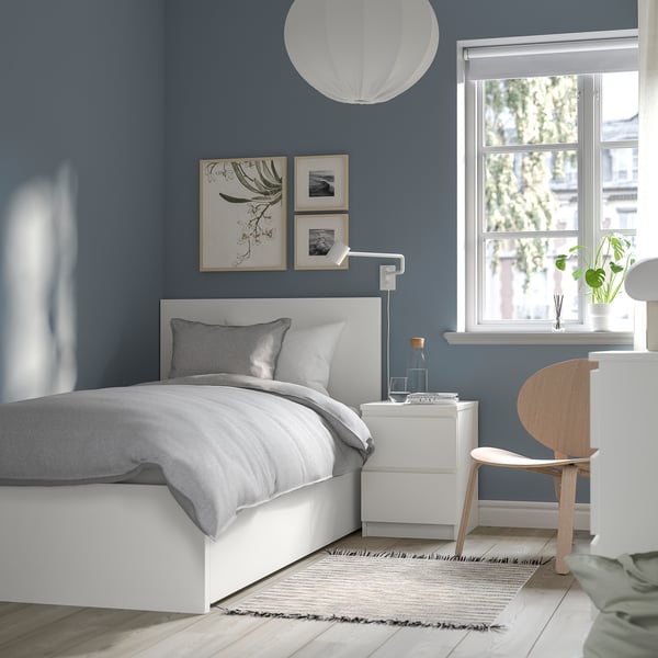 Camera da letto minimalista: letto MALM bianco centrato, lenzuola grigie, sedia in legno, tappeto bianco, parete blu, grande finestra.