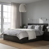 Camera da letto moderna e minimalista con letto nero, lenzuola grigie, grande finestra, legno scuro e pianta verde.