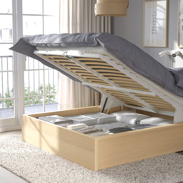 Un letto in legno chiaro con una base ribaltabile che rivela lo spazio di archiviazione con coperte e cuscini ben piegati. Il letto ha un piumino grigio sulla parte superiore.