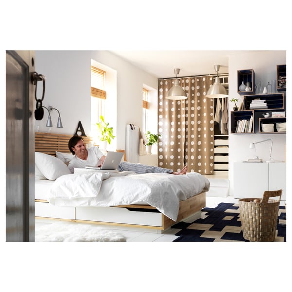 Una donna si rilassa su un letto di legno bianco con cassetti, usando un laptop. La stanza è dotata di una tenda a pois, di un cesto intrecciato e di scaffali con oggetti decorati.