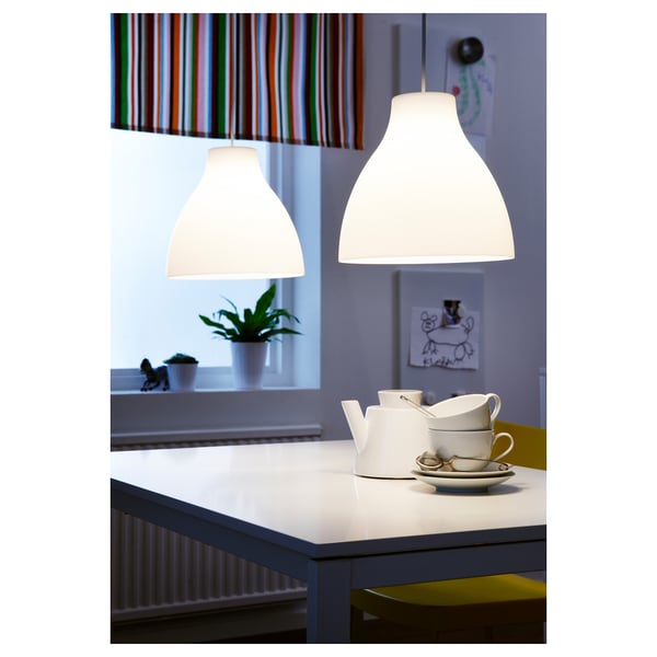 Due lampade a sospensione illuminano un tavolo da pranzo con teiera e tazze. Una tenda a strisce e una pianta sono sullo sfondo.