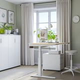 Configurazione dell'ufficio domestico con scrivania bianca MITTZON sit/stand con grande finestra, armadio bianco, orologio, piante e sedia.