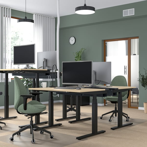 Ufficio moderno con scrivanie sit-stand MITTZON in legno nero e chiaro, sedie verdi, monitor e piante. Scrivanie più grandi di sedie e monitor.