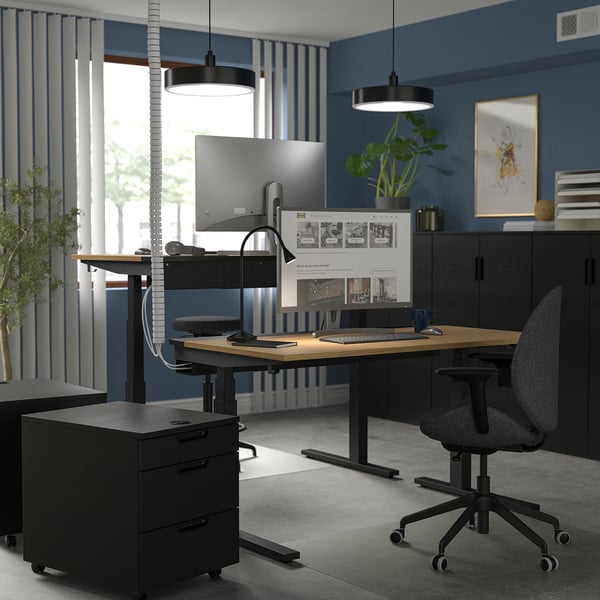 Moderno home office con scrivania sit/stand MITTZON, nero e legno, doppio monitor, sedia ergonomica, armadio nero, pareti blu, grande finestra, piante.