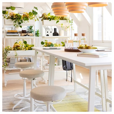 Spazio di lavoro luminoso e ben illuminato con tavoli, sgabelli e scaffali bianchi. Elementi di verde e arredamento esposti. Luce naturale dalle finestre.