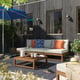 Moderno patio esterno con divano grigio NÄMMARÖ, cuscini arancioni e blu, tavolino in legno, ombrellone blu, piante e sfondo scuro della casa.