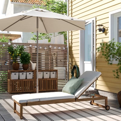 Cortile con grande lettino prendisole beige NÄMMARÖ, cuscino e ripostiglio in legno, piante e casa beige.