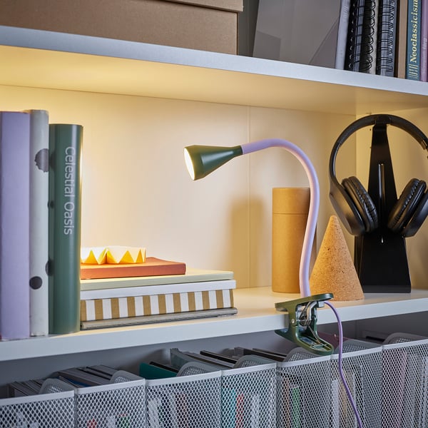 Libreria con lampada NÄVLINGE fissata sul bordo, libri luminosi. Sono visibili anche cuffie e varie pile di libri.
