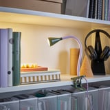 Libreria con lampada NÄVLINGE fissata sul bordo, libri luminosi. Sono visibili anche cuffie e varie pile di libri.