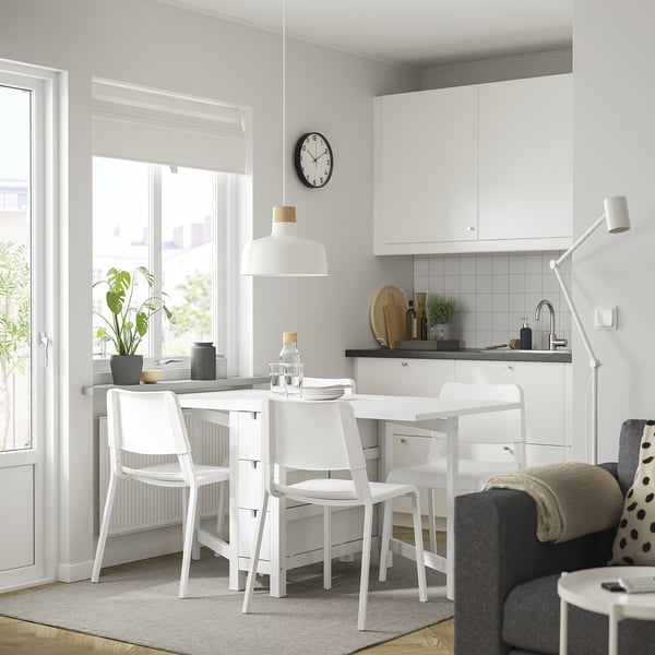 Cucina moderna con tavolo NORDEN e sedie impilabili. Il tavolo è dotato di cassetti, vicino alla finestra con lampada a sospensione. Armadi bianchi, piano di lavoro grigio e tappeto.